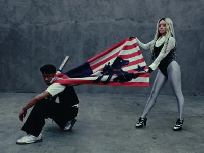 Two performers pose against a raw concrete backdrop in a stylized scene from PARTYOF2’s “PUNK B!TCH” visual. One crouches in monochrome streetwear and metallic sneakers while the other stands in platform heels, silver tights, and a layered black-and-white look. Between them, they stretch a custom red, white, and blue flag featuring a bold black graphic motif, creating a tense, theatrical composition that mirrors the track’s punk-meets-rage energy