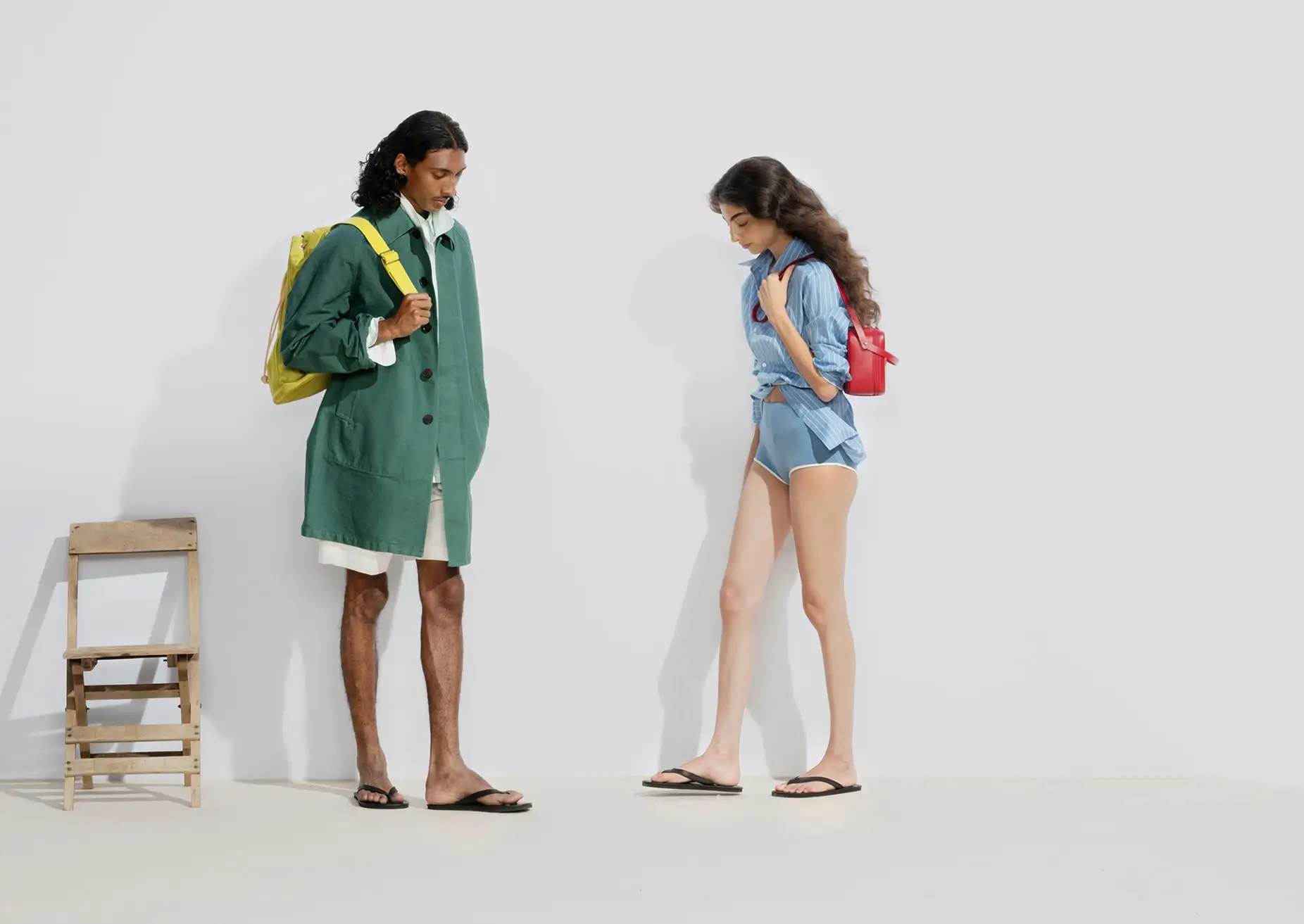 Two models wearing minimalist layered looks with oversized outerwear, striped shirting, flip-flops, and colorful backpacks against a pale studio backdrop in AURALEE Spring/Summer 2026 campaign imagery