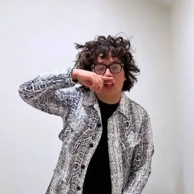 Casual portrait of a person with messy curly dark hair and round black glasses standing against a plain white wall. They wear a light gray snakeskin-pattern overshirt layered over a black top and pose playfully with a hand raised near their mouth, giving the image an informal, expressive, and candid feel