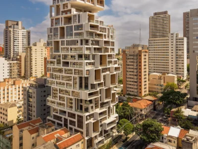 A towering contemporary residential high-rise rises above a dense urban neighborhood, defined by a dramatic stacked-cube architecture with oversized cantilevered volumes, recessed terraces, and a rigid grid of glass and concrete framing. The building’s sculptural silhouette contrasts against surrounding mid-century apartment blocks and low-rise homes with terracotta roofs, while bright afternoon sunlight and scattered clouds amplify the structure’s geometric depth and warm neutral tones
