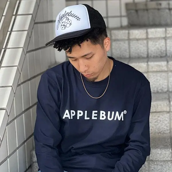 A young man sits on tiled outdoor steps, leaning slightly forward with his head tilted down in a contemplative pose. He wears a black-and-white trucker cap with a graphic logo, a navy long-sleeve shirt reading “APPLEBUM” in bold white lettering, and a thin gold chain necklace. His short curly hair peeks out from under the cap. The setting features light gray tiled walls and steps, creating a clean, urban backdrop with soft natural lighting