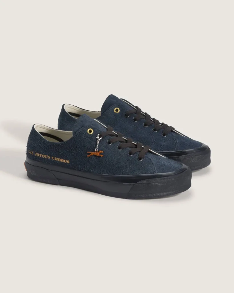 Dark navy suede low-top Vans shoe featuring a minimalist monochrome design, black sole unit and laces, gold “The Joyous Chorus” heel lettering, and a small metallic charm detail attached near the eyelets against a neutral studio backdrop
