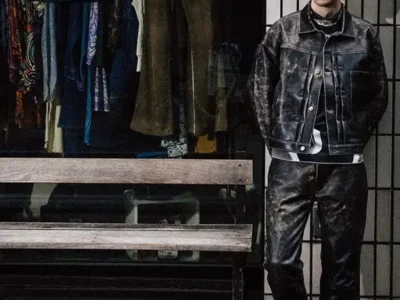 Model with short bleached hair wearing a distressed black leather jacket and matching leather pants, standing beside a storefront window filled with hanging garments in a dim urban streetscape
