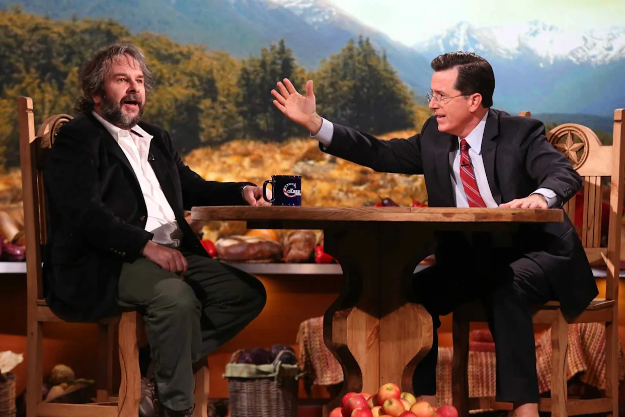 Two men sit across from each other at a rustic wooden talk-show set, engaged in animated conversation. One gestures with an outstretched hand while the other reacts mid-sentence. The backdrop features a scenic landscape of trees and snow-capped mountains, with warm studio lighting and seasonal table details creating a cozy, lodge-inspired atmosphere