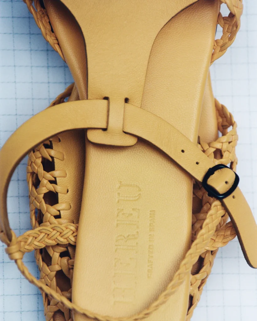 Close-up of woven leather sandal in muted camel tone with braided straps, adjustable buckle fastening, and embossed branding across the cushioned footbed on a grid-pattern backdrop