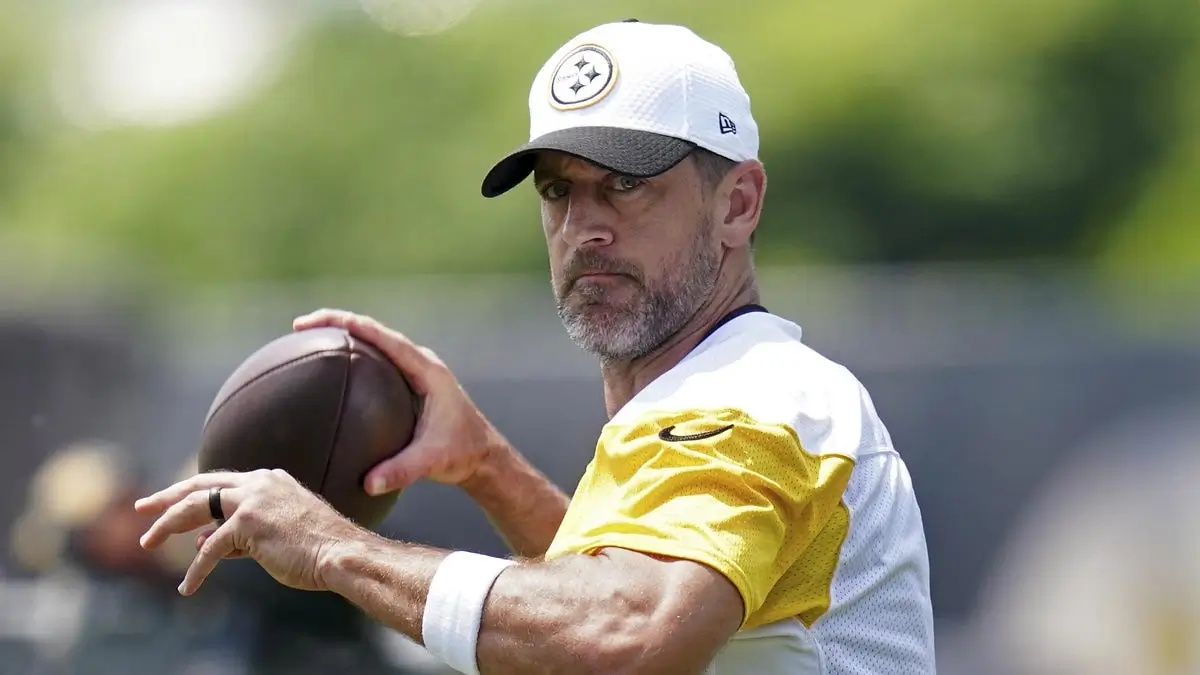 Close-up training field image of a veteran quarterback in Pittsburgh Steelers practice gear holding a football mid-throw. Wearing a white Steelers cap and yellow practice jersey, he looks intensely downfield during an outdoor session with a softly blurred green background