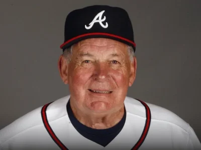 Portrait of legendary Bobby Cox wearing an Atlanta Braves cap and white Braves jersey, photographed against a dark neutral background. The longtime Hall of Fame manager appears calm and approachable, reflecting the steady leadership and warmth that defined his decades-long baseball legacy