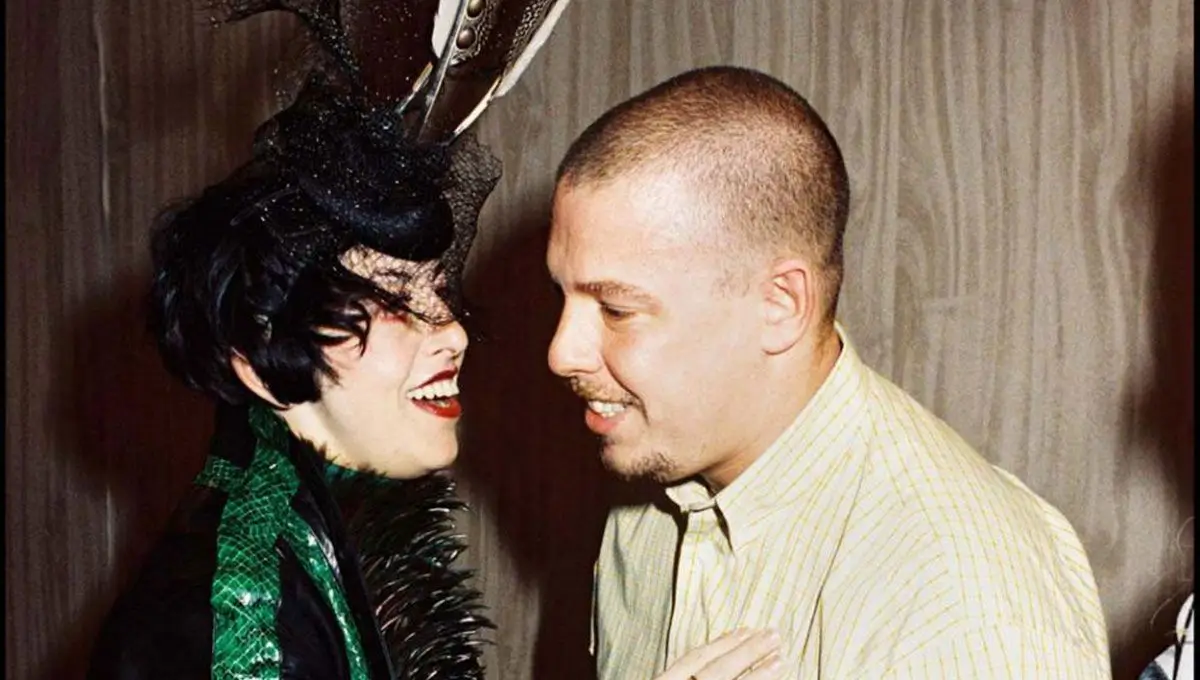 Isabella Blow and Alexander McQueen share an intimate backstage moment in this candid portrait, capturing the magnetic creative chemistry that defined one of fashion’s most legendary partnerships. Blow’s dramatic feathered headpiece, theatrical styling, and vivid personality contrast beautifully with McQueen’s understated tailoring and grounded presence, reflecting the collision of aristocratic eccentricity and working-class brilliance that fueled their transformative impact on 1990s and early-2000s fashion culture