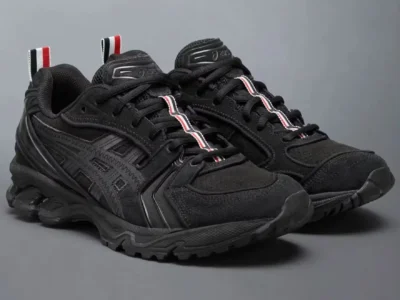 Black ASICS trail-inspired shoe with tonal layered overlays, chunky rugged sole, and red-white-navy striped pull tabs against a gray studio background