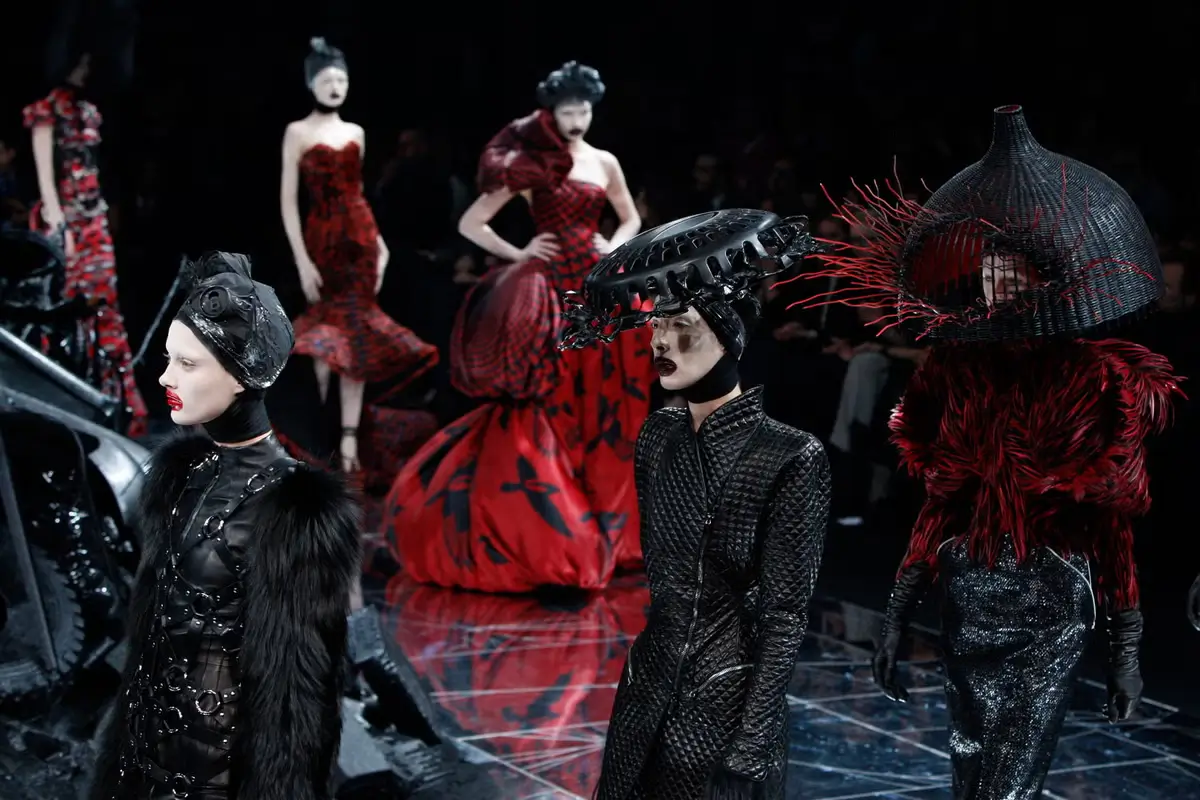 Models walk through a darkly theatrical Alexander McQueen runway presentation wearing sculptural silhouettes, dramatic headpieces, and intensely textured garments that blur the line between couture, performance art, and gothic fantasy. Dominated by black and crimson tones, the collection channels the emotional violence, romanticism, and avant-garde spectacle that defined Lee Alexander McQueen’s creative universe, where fashion became an immersive exploration of beauty, power, and psychological tension