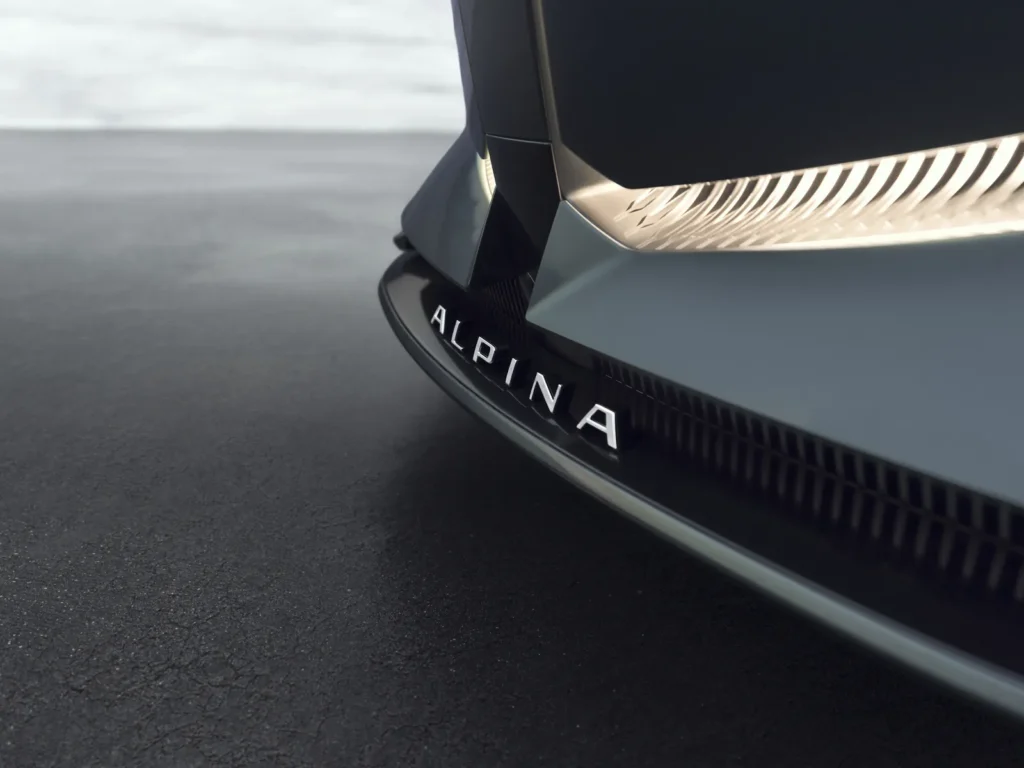 A cinematic close-up of the Vision BMW ALPINA’s illuminated front fascia, showcasing the concept’s sharply sculpted shark-nose geometry and integrated warm-white lighting signature. The polished ALPINA wordmark sits embedded within the lower intake, framed by dark metallic aero detailing and reflective black trim. Against the muted asphalt and overcast horizon, the composition emphasizes the brand’s restrained luxury language, where subtle surfacing and precision illumination replace overt aggression with architectural sophistication