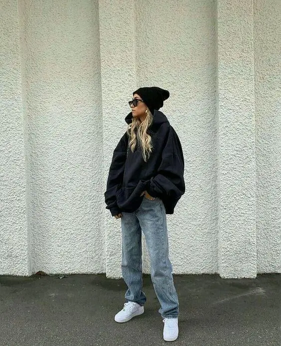 Person standing against a textured wall wearing an oversized black hoodie, light-wash loose jeans, white sneakers, and a black beanie with sunglasses, styled in a relaxed streetwear look