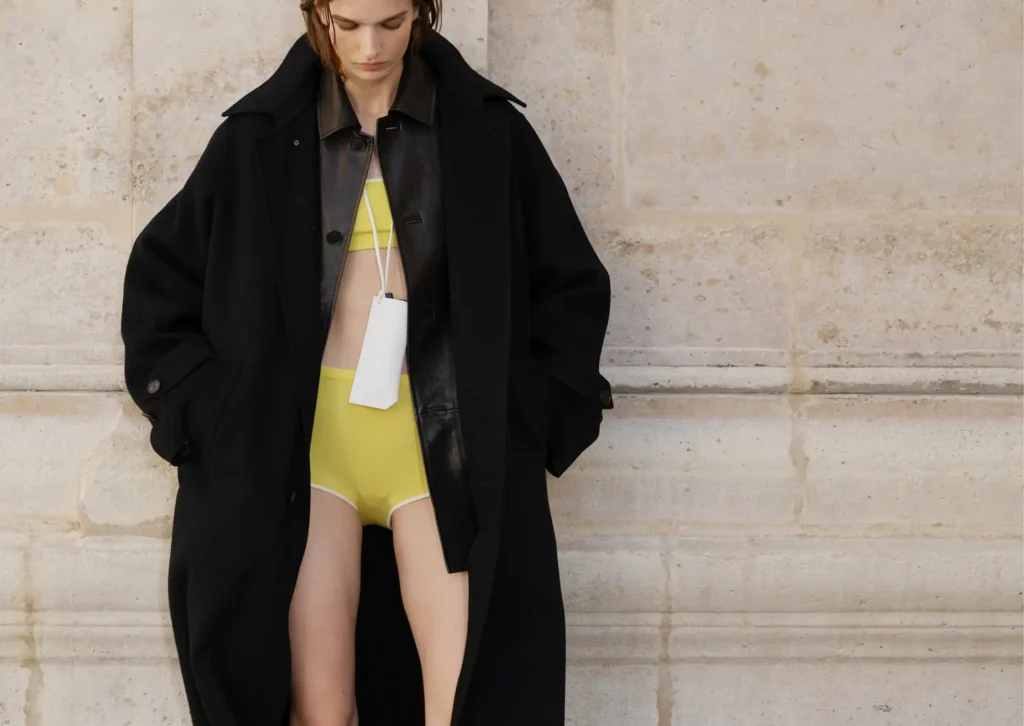 Model wearing an oversized black overcoat layered over a dark leather jacket and bright yellow two-piece set against a pale stone wall during AURALEE Spring/Summer 2026 campaign