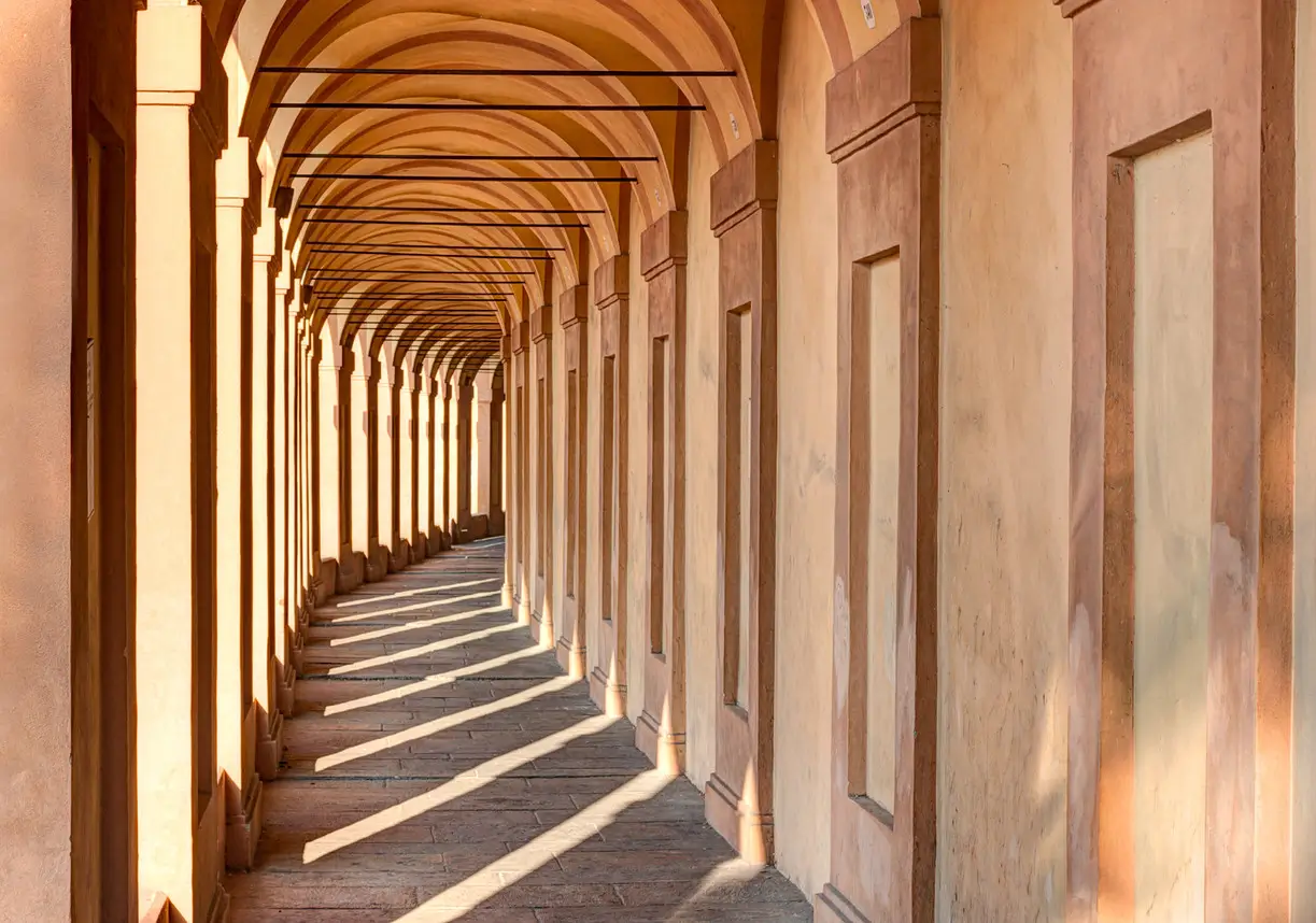 Historic Bologna portici corridor featuring repeating sandstone arches, warm terracotta tones, and dramatic afternoon light casting long geometric silhouettes across the stone walkway