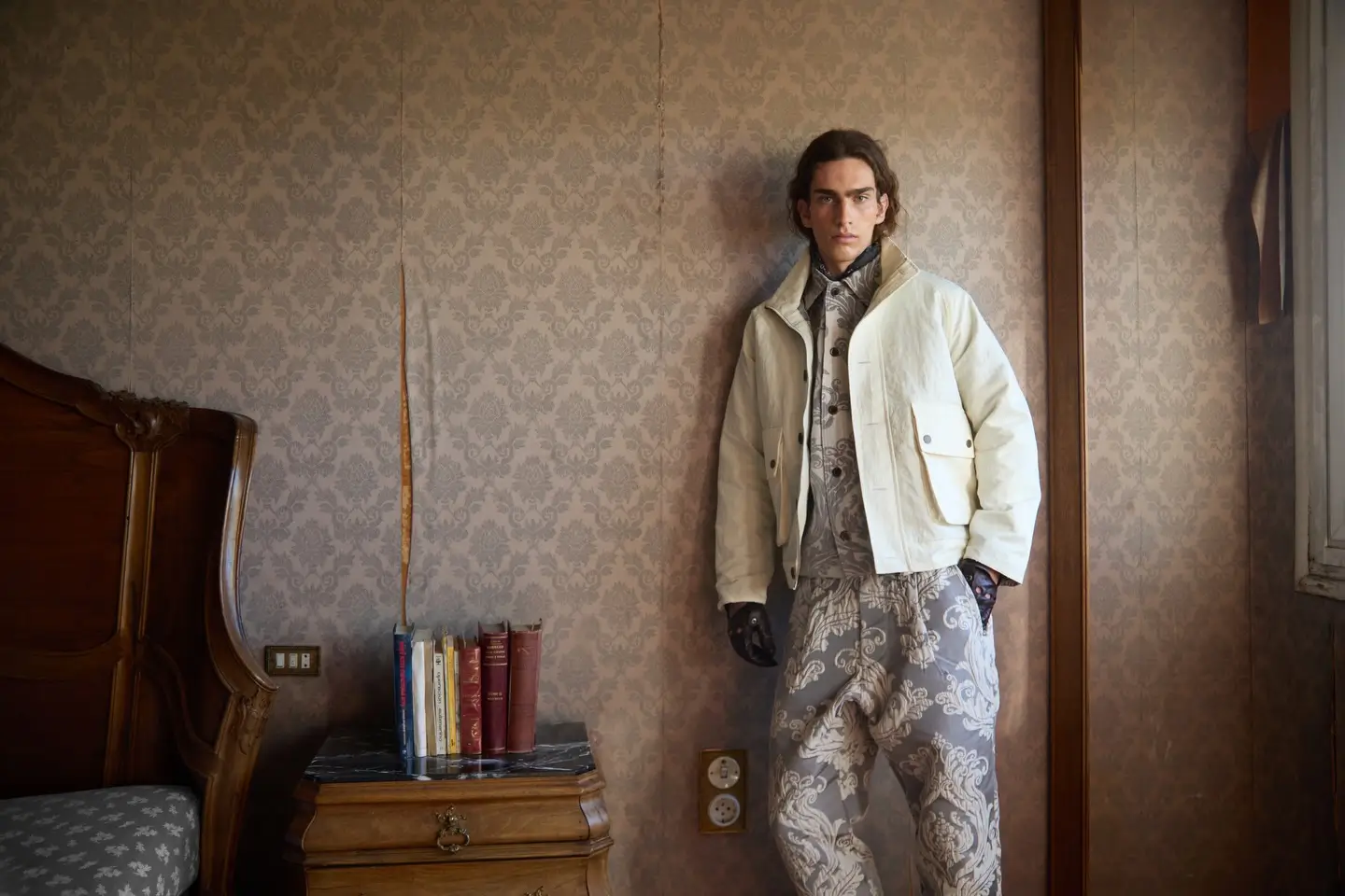 A model stands against faded damask wallpaper inside a weathered vintage room, wearing an editorial ensemble that merges romantic tailoring with utilitarian outerwear. The look pairs a cropped ivory jacket with ornate monochromatic patterned layers and matching trousers, while dark leather gloves sharpen the silhouette with a subtle gothic tension. Antique wooden furniture, stacked books, and peeling interiors reinforce the atmosphere of quiet decay, creating a cinematic fashion composition that feels both aristocratic and hauntingly contemporary