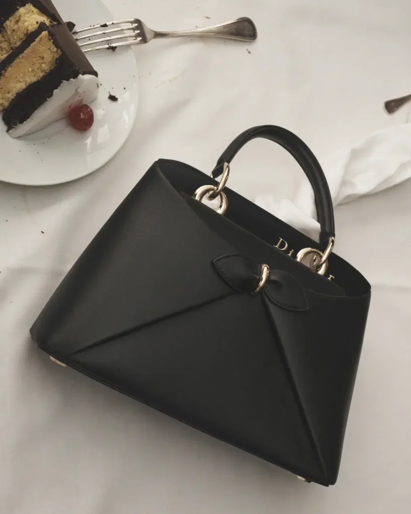 An overhead view of a white café table featuring a structured black leather handbag with a sculptural folded front and a subtle bow-like detail, accented by gold hardware and a curved top handle. Nearby, a partially eaten slice of layered cake sits on a white plate with a fork resting above it, with a small red cherry garnish and scattered crumbs adding a casual, lived-in touch. A crumpled napkin lies beside the bag, softening the composition. The clean white surface and minimal palette contrast with the rich texture of the bag, creating a refined yet relaxed café still-life