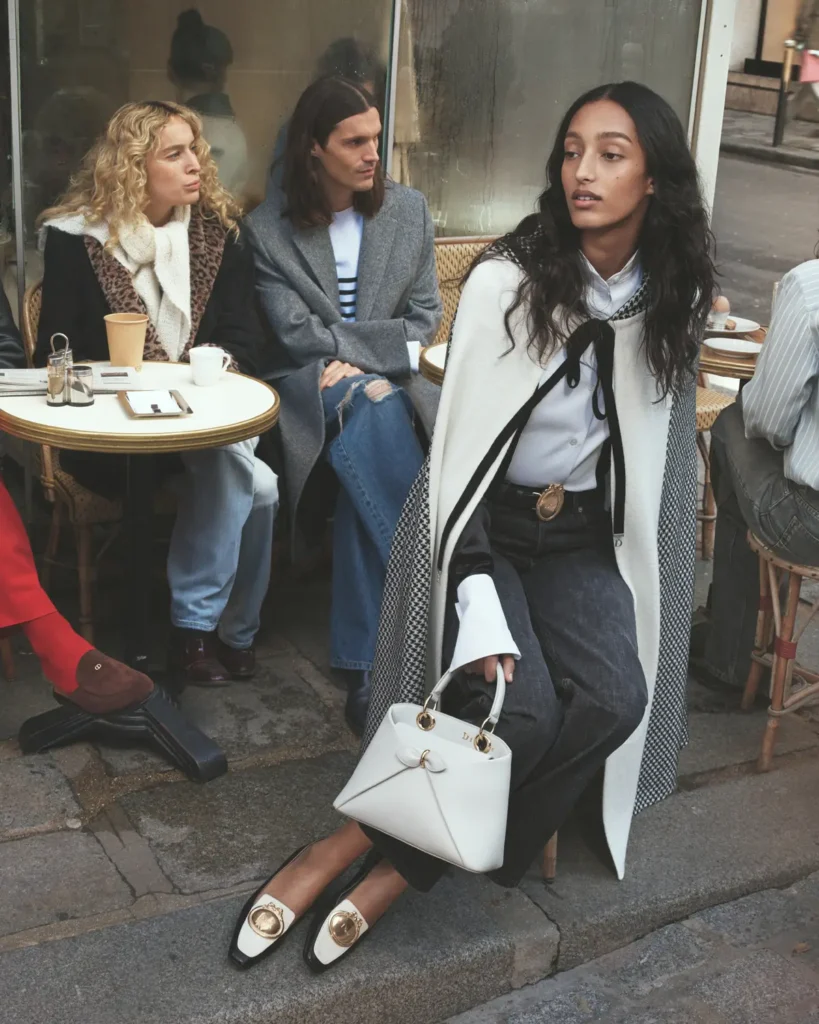 A woman sits poised at the edge of a sidewalk café, turned slightly as if caught mid-thought, holding a structured white handbag with gold hardware. She wears a crisp white blouse layered under a draped, tailored cape with contrasting textures, paired with dark trousers and pointed flats adorned with bold metallic accents. Behind her, two seated figures at a small round café table—one in a leopard-print coat with a scarf, the other in a gray overcoat and denim—appear engaged in quiet conversation, adding a sense of everyday Parisian life. The scene is framed by a glass window with soft reflections, muted street tones, and natural light, creating a candid yet composed moment that blends street style with refined elegance