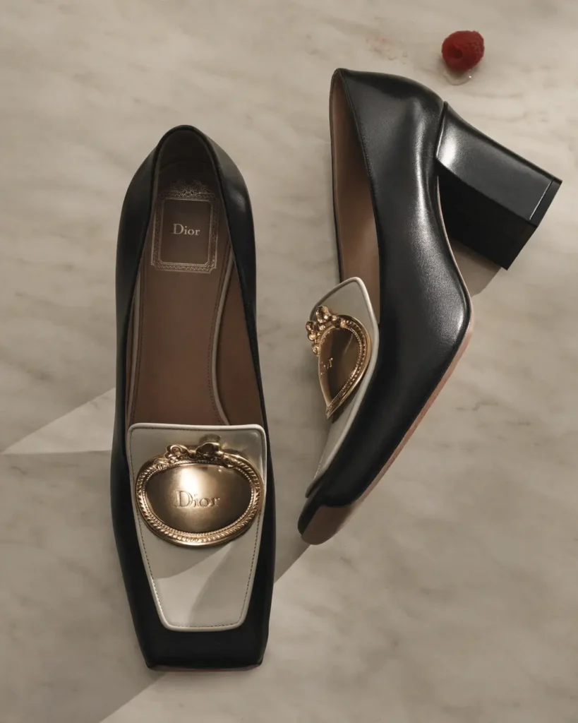 An overhead still-life of a pair of black leather heeled loafers set against a pale marble surface. The shoes feature a sculpted, slightly squared silhouette with a low block heel and a contrasting light panel at the vamp, each adorned with an ornate oval gold plaque engraved with Dior branding. One shoe lies flat while the other is angled to reveal its profile, highlighting the smooth leather finish and structured form. A single raspberry nearby adds a subtle pop of color, reinforcing the refined, café-inspired aesthetic