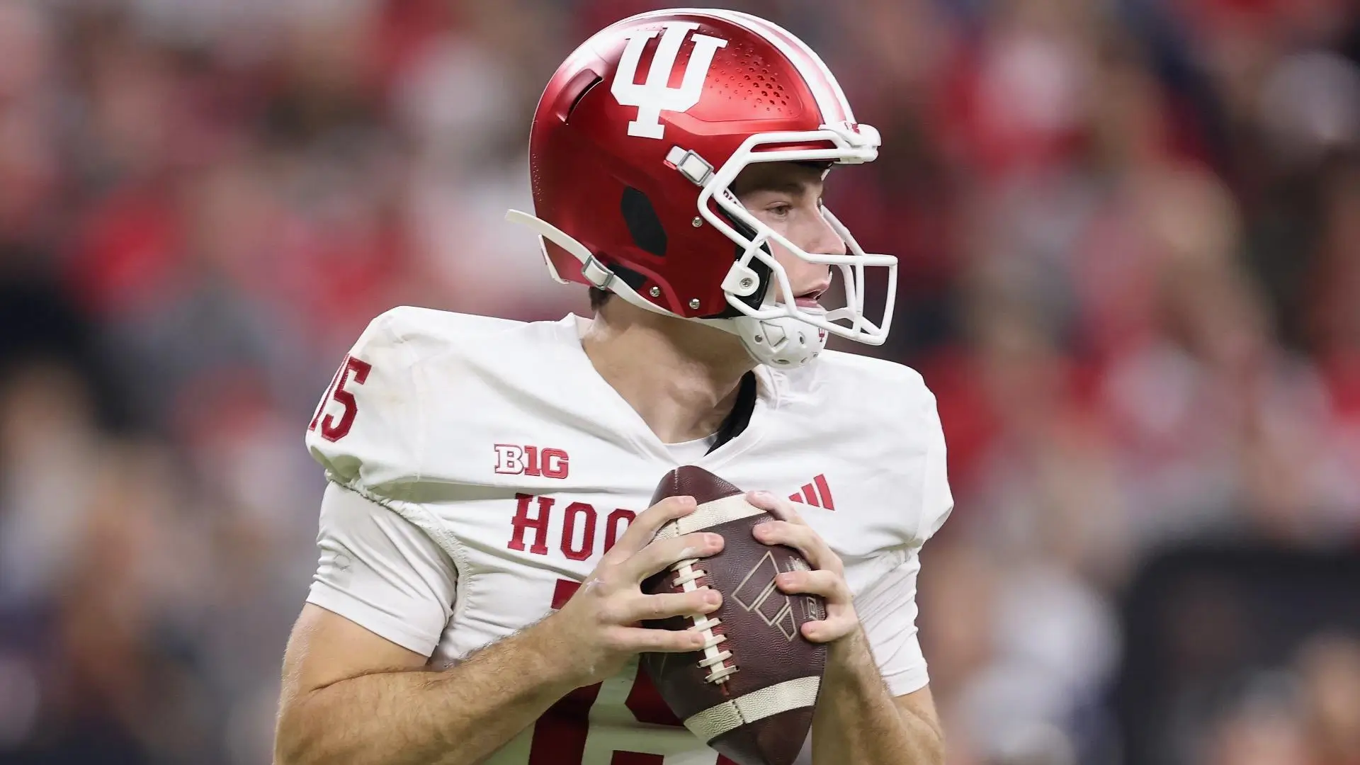 Quarterback in a white Indiana Hoosiers uniform and red helmet (#15) holds the football with both hands, looking right to pass during a game, with a blurred stadium crowd in the background