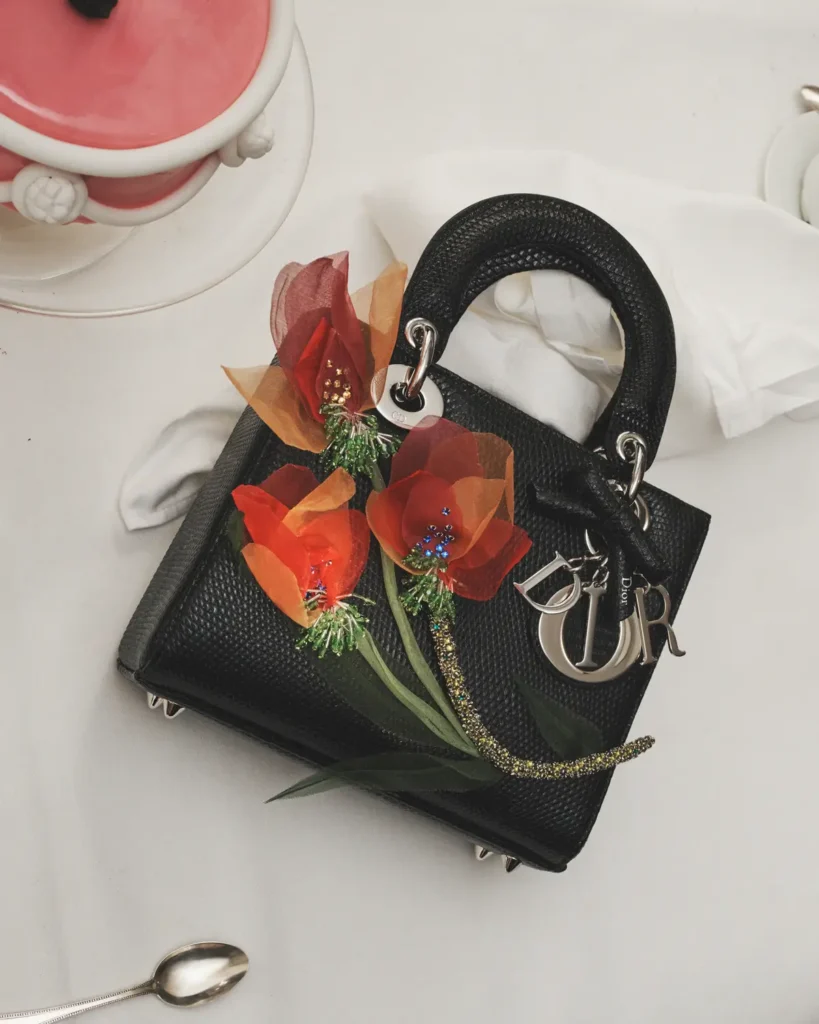 An overhead still-life of a black textured leather handbag set against a clean white café table, its structured silhouette accented by silver Dior letter charms and rounded handles. Draped across the bag are vivid, translucent floral embellishments in red and burnt orange tones, with delicate beaded centers that introduce a handcrafted, almost couture-like contrast to the pebbled leather. A crumpled napkin and a spoon rest nearby, while the edge of a pink-glazed dessert enters the frame, softening the composition. The scene balances precision and spontaneity—refined luxury interrupted by small, lived-in gestures