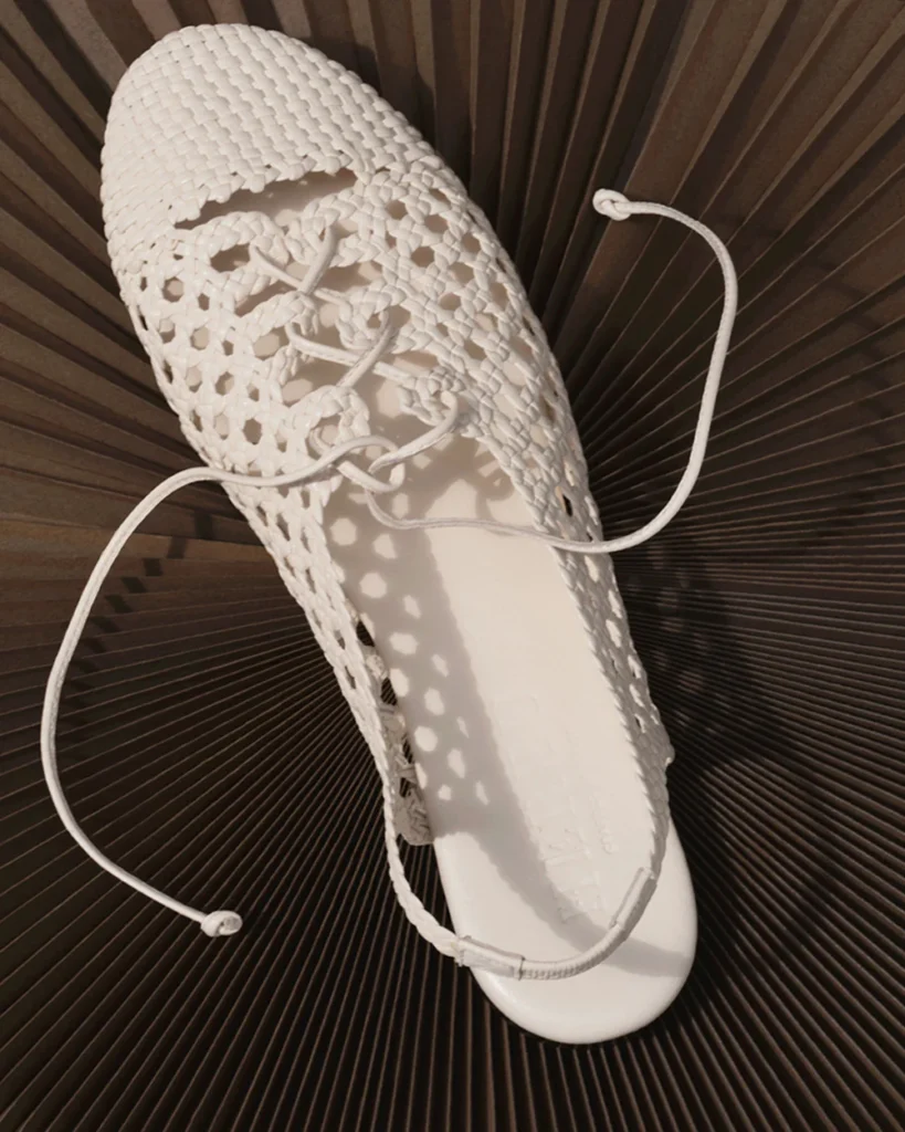 White woven lace-up sandal with intricate open-knit construction, elongated cord ties, and glossy molded footbed, styled against a dark pleated backdrop with dramatic flow