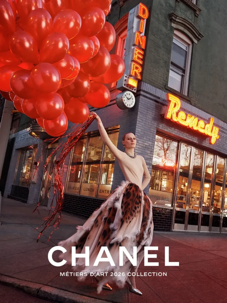 Nighttime CHANEL campaign portrait outside a neon diner with a model holding oversized red balloons while wearing a dramatic feather-trimmed animal-print skirt