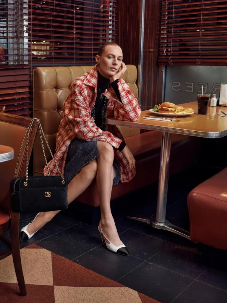 Editorial diner scene from the CHANEL Métiers d’Art Collection showing a model seated in a vintage booth wearing a red-and-white checked coat, black turtleneck layers, two-tone slingback heels, and a classic quilted Chanel shoulder bag