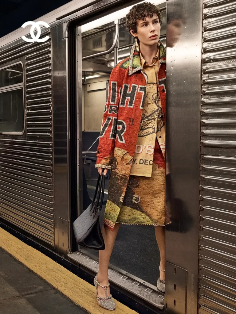 Subway platform fashion campaign featuring a model stepping from a train in layered graphic tailoring and textured separates, styled with crystal-embellished shoes and an oversized black Chanel tote