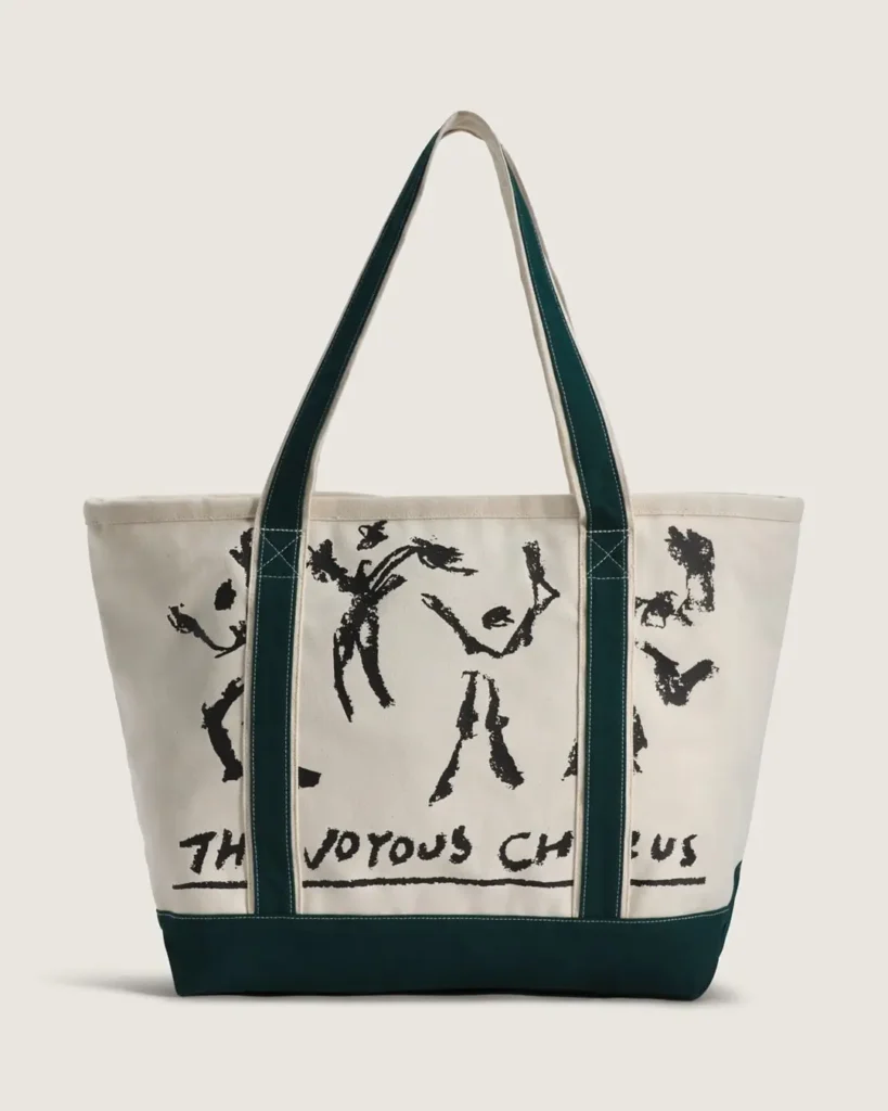Oversized canvas tote bag with deep green trim and shoulder straps, featuring large hand-drawn abstract figures and “The Joyous Chorus” text printed across the natural canvas body in a raw artistic style against a clean neutral background