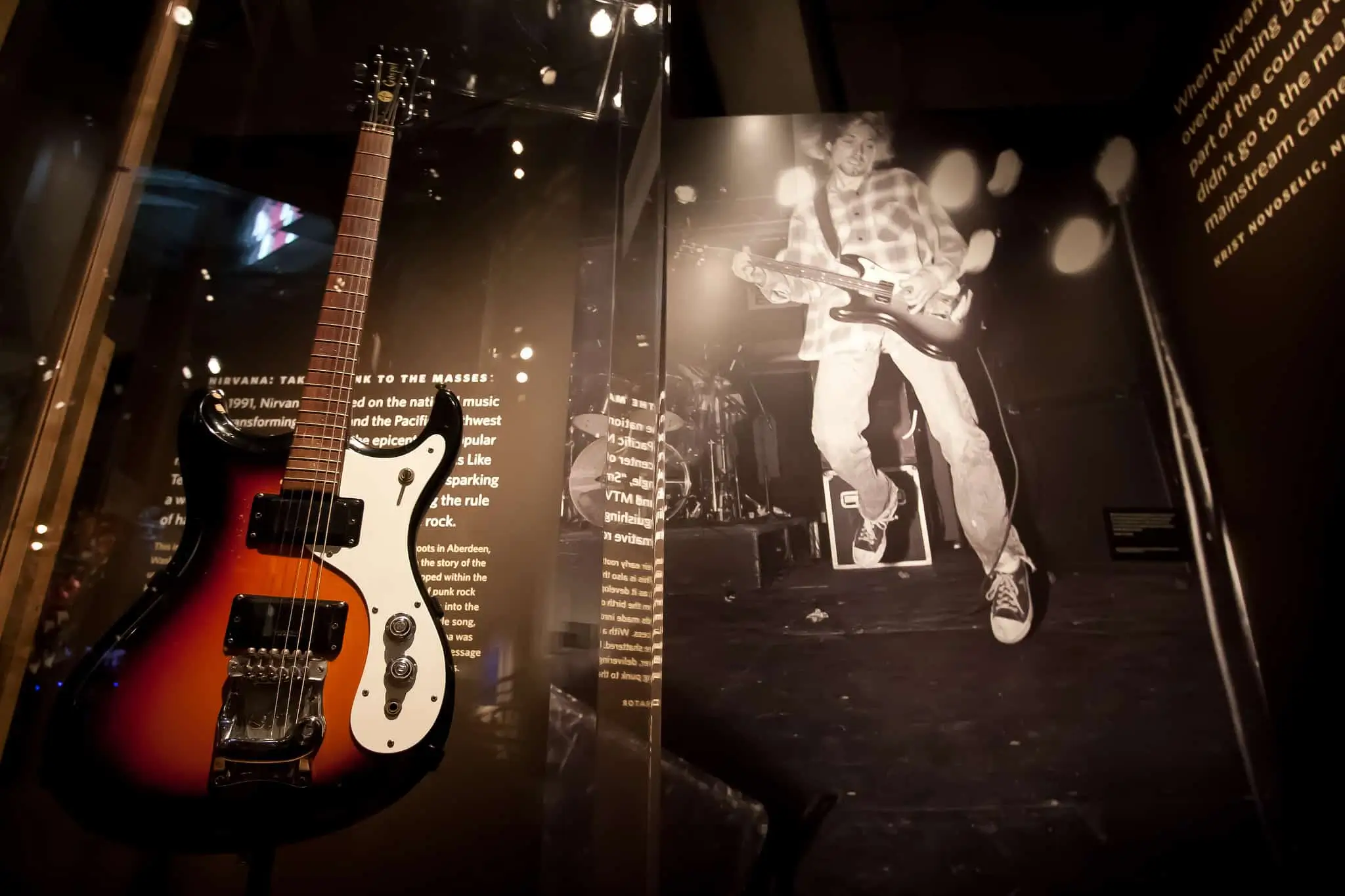 Dimly lit exhibition display inside the GRAMMY Museum featuring a vintage sunburst electric guitar alongside a large black-and-white performance image of Kurt Cobain, capturing the raw energy and cultural legacy of the grunge era through archival music memorabilia