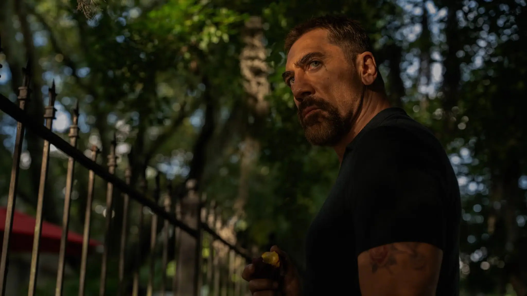 A moody cinematic portrait of a man standing beside a wrought-iron fence beneath dense trees draped in hanging moss. Wearing a fitted black T-shirt, he looks off-frame with a guarded, intense expression as warm evening light cuts across his face and arm, creating a suspenseful Southern gothic atmosphere