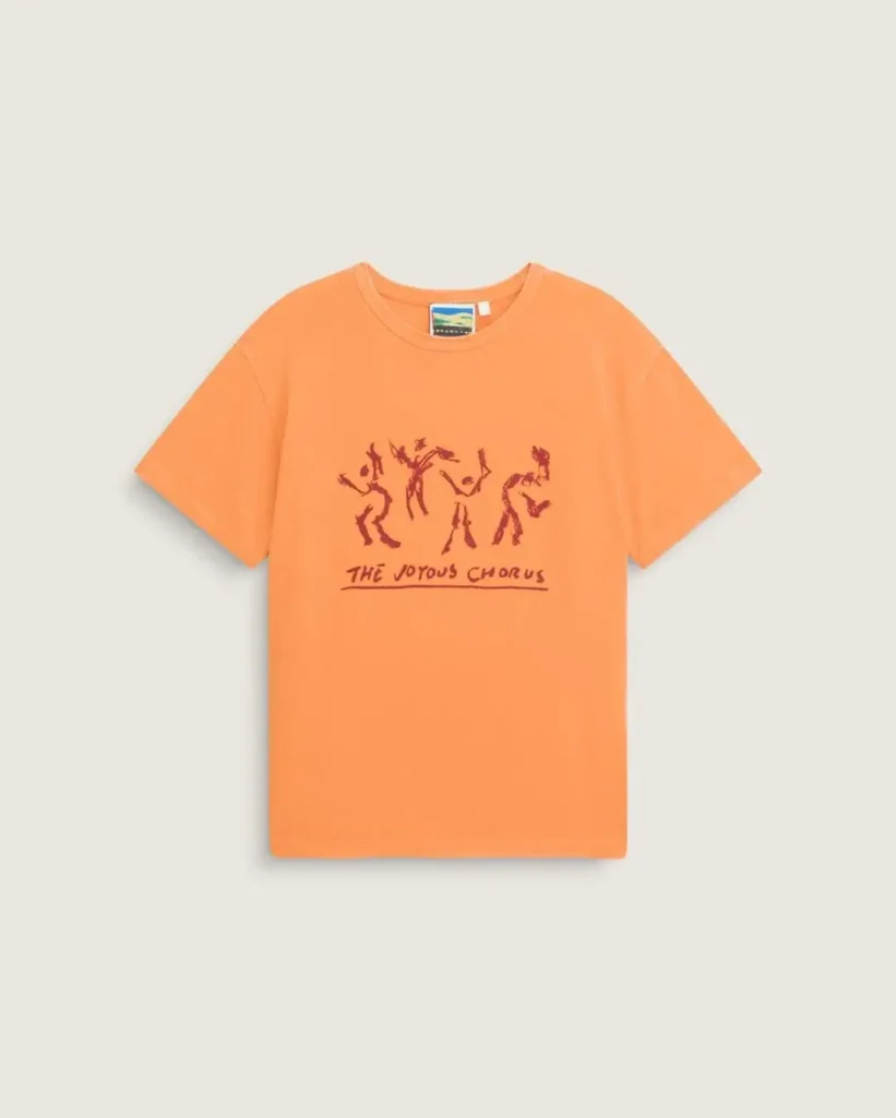 Soft orange graphic T-shirt featuring hand-drawn dancing figures and “The Joyous Chorus” lettering across the chest, designed with a vintage-washed finish and relaxed silhouette against a minimal neutral background