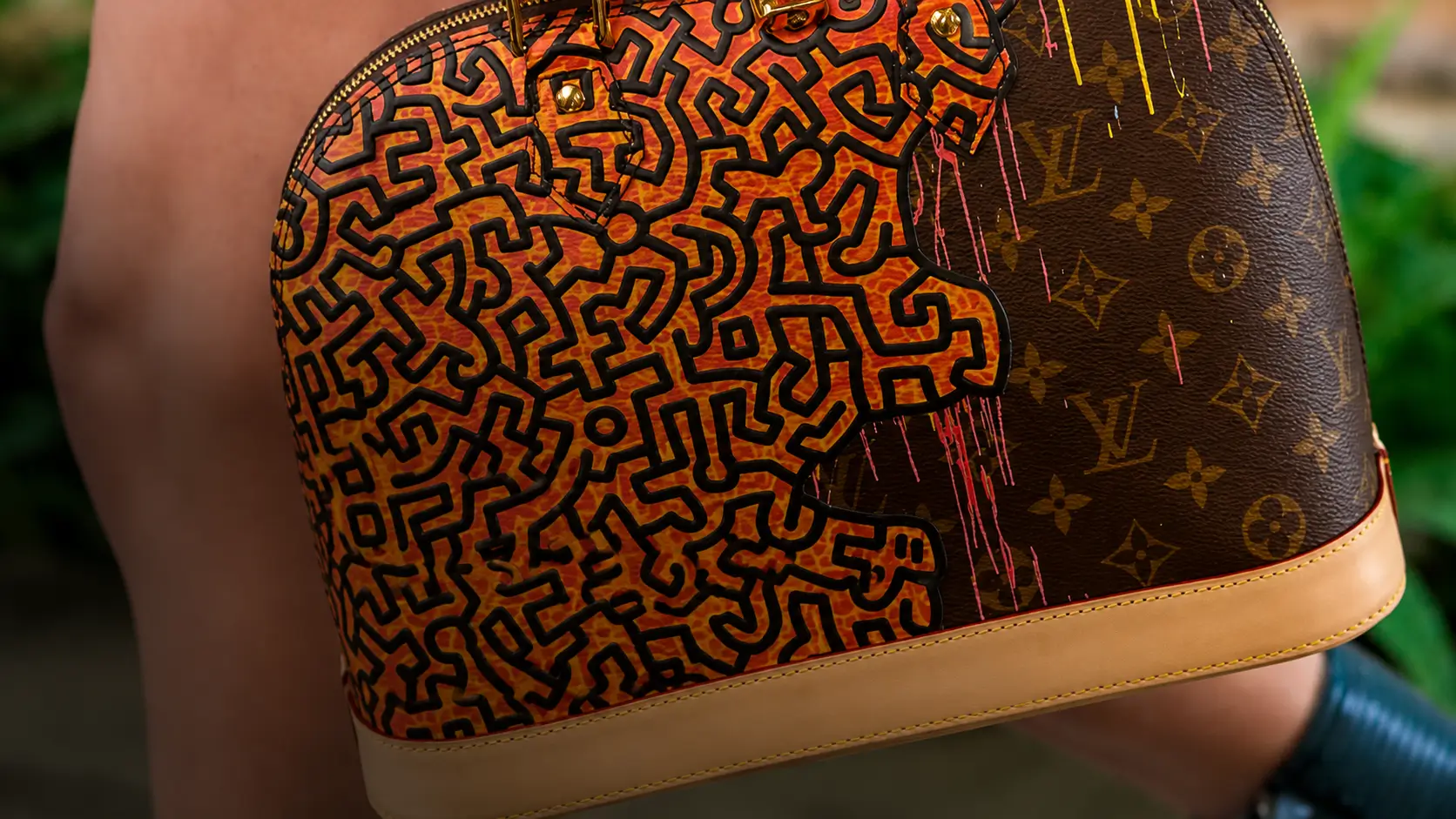 Close-up detail of a haute handbag featuring a vivid orange-and-black abstract line artwork layered against classic monogram canvas, finished with natural leather trim and gold hardware