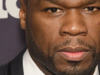 Close-up portrait of a man in formal attire photographed against an event backdrop. The tightly framed image emphasizes direct eye contact, textured lighting, and a composed expression, creating a polished red-carpet editorial aesthetic