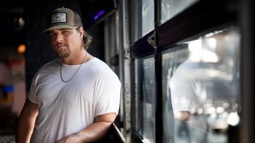 A moody portrait of country artist Gavin Adcock standing beside a reflective industrial-style window in a dimly lit interior. He wears a fitted white T-shirt, a gold chain necklace, and a brown trucker cap with a waterfowl patch, reinforcing the rugged Southern aesthetic tied closely to his public image. Soft natural light cuts across his face and shoulders while the blurred reflection in the glass adds depth and atmosphere. The composition balances modern country imagery with a stripped-back realism, emphasizing Adcock’s laid-back yet confrontational presence associated with his bro-country persona
