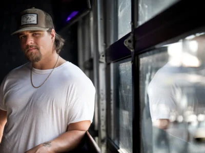 A moody portrait of country artist Gavin Adcock standing beside a reflective industrial-style window in a dimly lit interior. He wears a fitted white T-shirt, a gold chain necklace, and a brown trucker cap with a waterfowl patch, reinforcing the rugged Southern aesthetic tied closely to his public image. Soft natural light cuts across his face and shoulders while the blurred reflection in the glass adds depth and atmosphere. The composition balances modern country imagery with a stripped-back realism, emphasizing Adcock’s laid-back yet confrontational presence associated with his bro-country persona