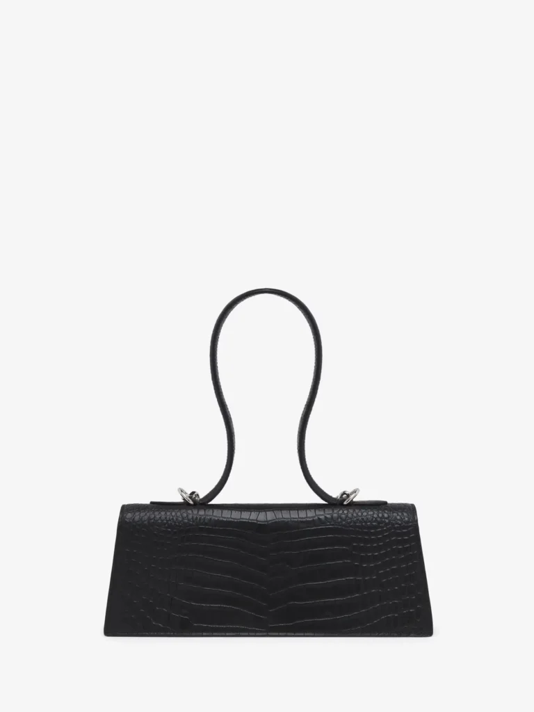 A black leather mini handbag presented front-on against a clean white background, emphasizing its elongated, structured silhouette. The body is low and horizontal, with a gently curved top edge and a firm, architectural base. The leather features a glossy crocodile-embossed finish, with symmetrical scale patterning that subtly radiates from the center, adding visual rhythm and texture. A distinctive, sculptural top handle rises in a smooth, continuous loop, creating an almost fluid, organic shape that contrasts with the bag’s rigid body. The handle is attached via small silver-tone metal rings on either side, introducing a minimal hardware accent without disrupting the design’s restraint. The overall composition feels balanced and intentional—sleek, compact, and quietly expressive through proportion and material rather than overt detailing