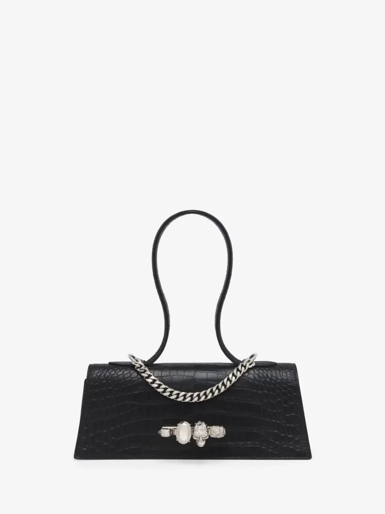 A black crocodile-embossed leather mini handbag presented front-on against a clean white background, defined by its elongated, low-profile silhouette and structured form. The glossy finish highlights the naturalistic scale pattern, which flows symmetrically across the body, giving the piece a refined, almost architectural surface texture. The sculptural top handle rises in a smooth, continuous loop, maintaining the bag’s minimalist foundation. Just below it, a silver-tone chain drapes across the upper front, introducing a layered, jewelry-like detail that adds movement and contrast against the dark leather. At the center of the bag sits a statement clasp composed of clustered, gem-like elements in polished silver tones. The hardware reads as both functional and ornamental—resembling a piece of fine jewelry embedded into the design—bringing a focal point to an otherwise restrained composition. The overall effect balances structure with embellishment, merging sharp proportions with subtle opulence