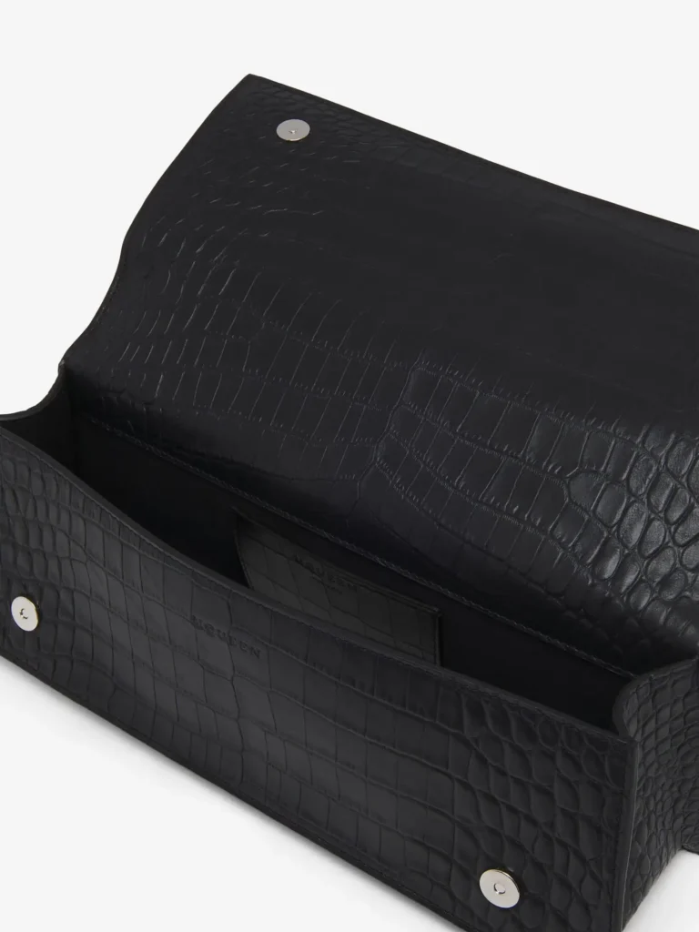 An open view of the black crocodile-embossed leather handbag, revealing its structured interior and construction. The flap is lifted to expose the underside, which carries the same glossy, textured finish, with a discreet circular magnetic snap closure set into the leather. The interior compartment appears compact yet neatly defined, maintaining the bag’s architectural rigidity. Inside, a subtle stamped logo is visible along the inner panel, reinforcing its luxury positioning without overt branding. The lining and internal surfaces continue the tonal black palette, emphasizing cohesion and restraint. The edges are cleanly finished, and the gusseted sides show how the bag expands slightly to accommodate essentials while retaining its sharp silhouette. The overall impression is one of precision craftsmanship—where exterior refinement extends seamlessly into the interior detailing