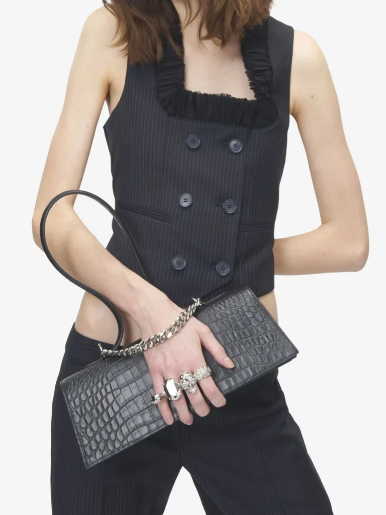 A styling shot of the black crocodile-embossed leather mini handbag worn against a clean white background, offering scale and context. The bag is held at the waist by a model dressed in a sharply tailored black pinstripe vest and matching trousers, creating a cohesive, monochrome look. The handbag’s elongated, structured silhouette contrasts with the fluidity of the body, while the sculptural looped handle curves upward in a soft, continuous arc. A silver-tone chain drapes across the top edge, adding a layered metallic accent. The statement clasp—composed of clustered, jewel-like elements—sits at the center front, aligning visually with the model’s hand, which is adorned with bold, oversized rings that echo the hardware’s reflective finish. The crocodile-embossed texture catches light subtly, reinforcing the bag’s refined surface detail without overpowering the composition. Against the tailored pinstripe ensemble, the piece reads as both sharp and decorative—balancing structure, ornament, and proportion in a distinctly modern, fashion-forward context