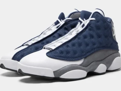 Air Jordan 13 shoe in a white, navy, and grey colorway featuring textured navy mesh overlays, smooth white leather uppers, grey suede mudguards, and signature holographic ankle detailing, shown in an angled studio product shot against a light grey background