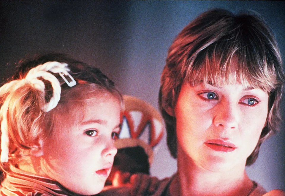 Softly lit cinematic close-up of an adult woman holding a young child while both look off frame with quiet concern and wonder. Warm pink and orange light washes across their faces, creating an intimate, nostalgic atmosphere. The child wears light-colored hair accessories and a striped top, while the woman’s expression suggests tenderness mixed with apprehension, reinforcing the scene’s emotional and protective tone