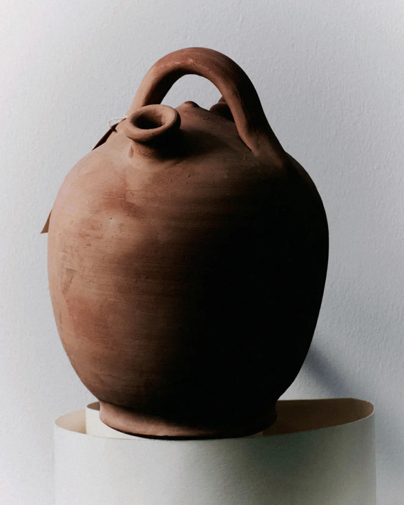 Rust-toned hand-thrown ceramic vessel with rounded body, sculptural loop handle, and narrow spout opening, displayed on a minimal pedestal under soft directional lighting