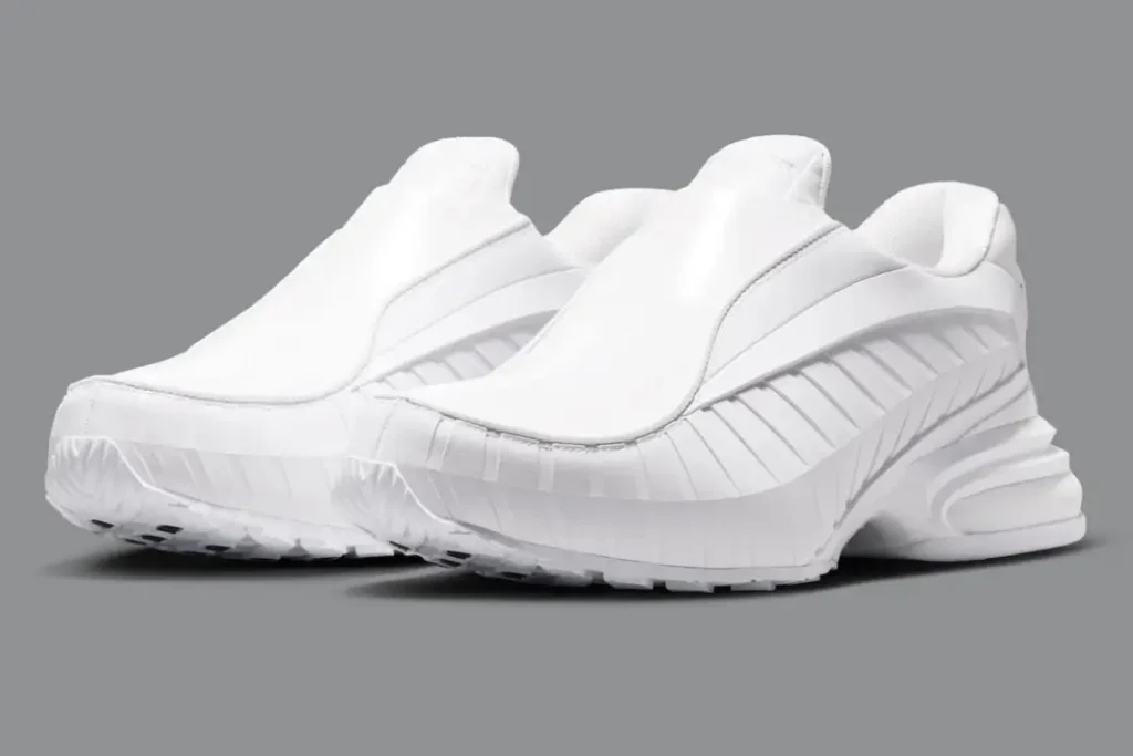 The all-white COMME des GARÇONS HOMME PLUS x Nike Air Max Dolce is displayed against a soft gray backdrop, emphasizing its sculptural slip-on form and futuristic luxury aesthetic. The laceless silhouette features smooth matte and glossy leather paneling with layered ribbed sidewalls that flow toward the exaggerated heel structure. Tonal detailing throughout the upper and sole creates a minimalist monochromatic appearance, while the chunky Air Max-inspired rear cushioning and aggressive outsole maintain the sneaker’s performance-rooted DNA beneath its refined, avant-garde presentation