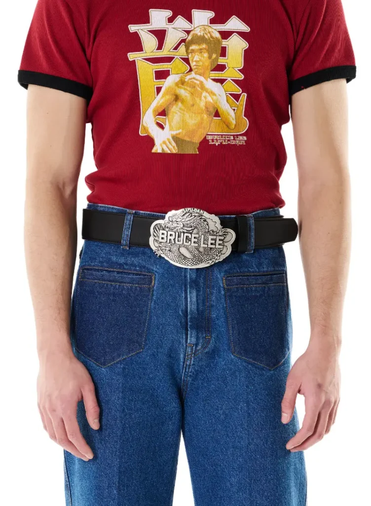 A close-up look from the LỰU ĐẠN x Bruce Lee collaboration showcases a fitted red ringer tee featuring archival-style Bruce Lee artwork layered over bold Chinese character graphics in yellow and cream tones. Styled with relaxed blue denim, the outfit is anchored by a black leather belt finished with an oversized silver “Bruce Lee” dragon buckle engraved with intricate detailing, channeling the capsule’s fusion of martial arts mythology, retro Americana, and rebellious luxury streetwear energy