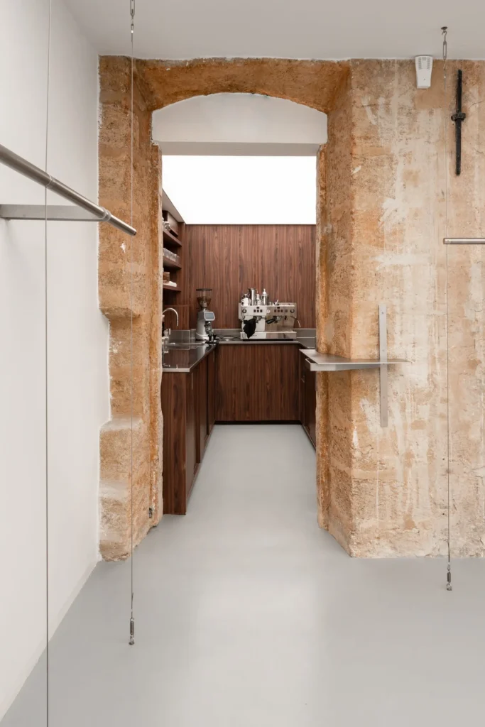 Minimal interior passage framed by an exposed limestone arch leading into a wood-clad café workspace, featuring stainless steel counters, espresso equipment, and suspended metal rails within a clean, gallery-like retail setting