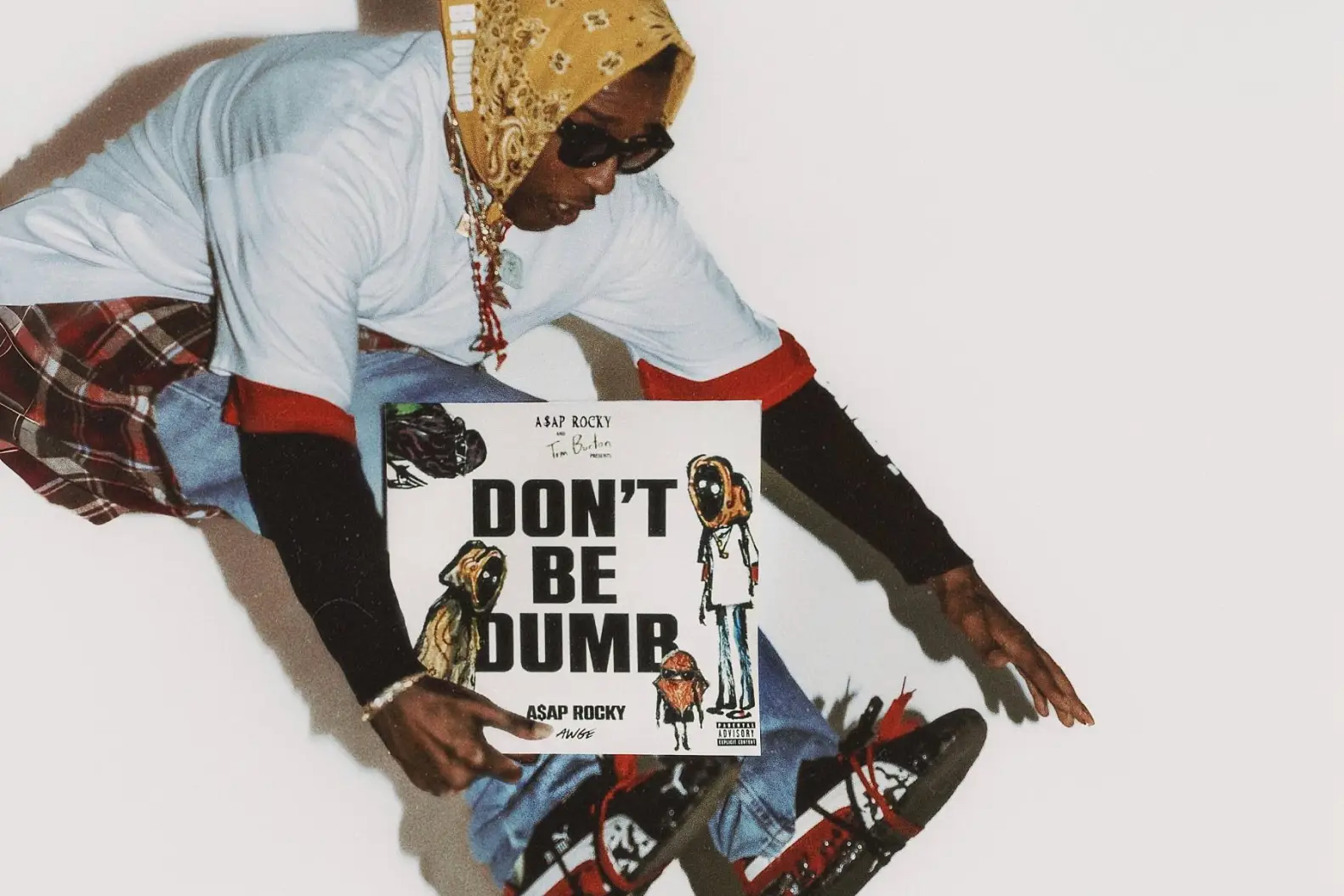 A grainy, editorial-style portrait of A$AP Rocky crouched against a stark white backdrop while holding the Don’t Be Dumb album artwork illustrated with Tim Burton-inspired characters. Rocky layers oversized streetwear with a yellow paisley headscarf, dark sunglasses, stacked jewelry, and black-red sneakers, reinforcing the album’s fractured alter-ego aesthetic through a raw, lo-fi view composition