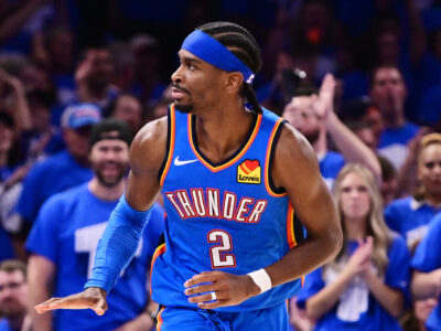 Shai Gilgeous-Alexander in a blue Oklahoma City Thunder uniform during a playoff game, moving across the court with a composed expression as a blurred sea of Thunder fans in blue fills the arena behind him