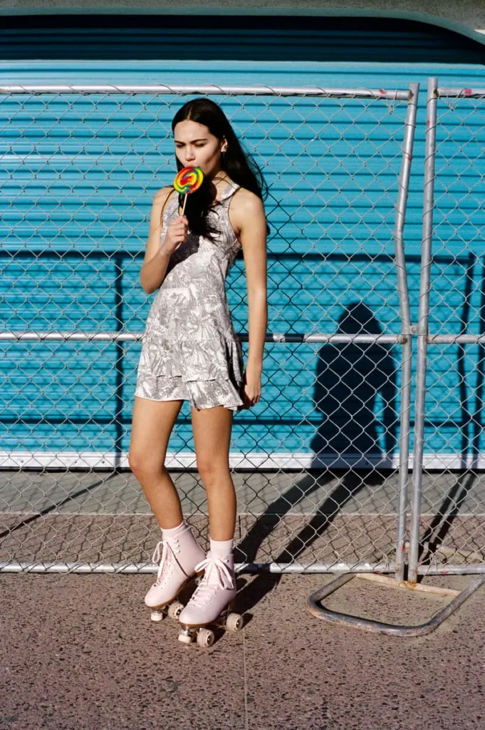 Sun-bleached innocence with a bite. She glides through her own constructed Eden — apple-print dress, roller skates, and a lollipop that feels like temptation in motion. Post-internet paradise, slightly sticky, perfectly off
