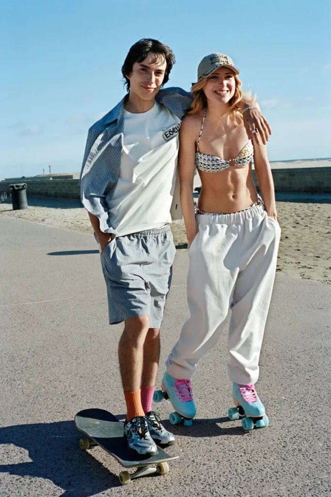Shoreline scripture. She stands serene in delicate patterned bikini, he owns the frame in oversized apple-print shorts — two modern originals, barefoot where the Pacific kisses their ankles. Golden hour has never felt more biblical… or more unbothered